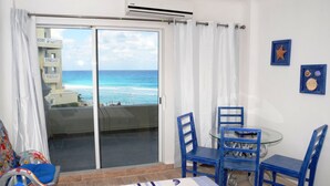 A room with a bed, a glass table with blue chairs, a view of the ocean through a sliding door, and a ceiling fan. - Amigo Rental (Cancun)