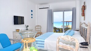 A hotel room with a bed, a blue sofa, a glass table with decorative items, a television, and a view of the beach. - Amigo Rental (Cancun)