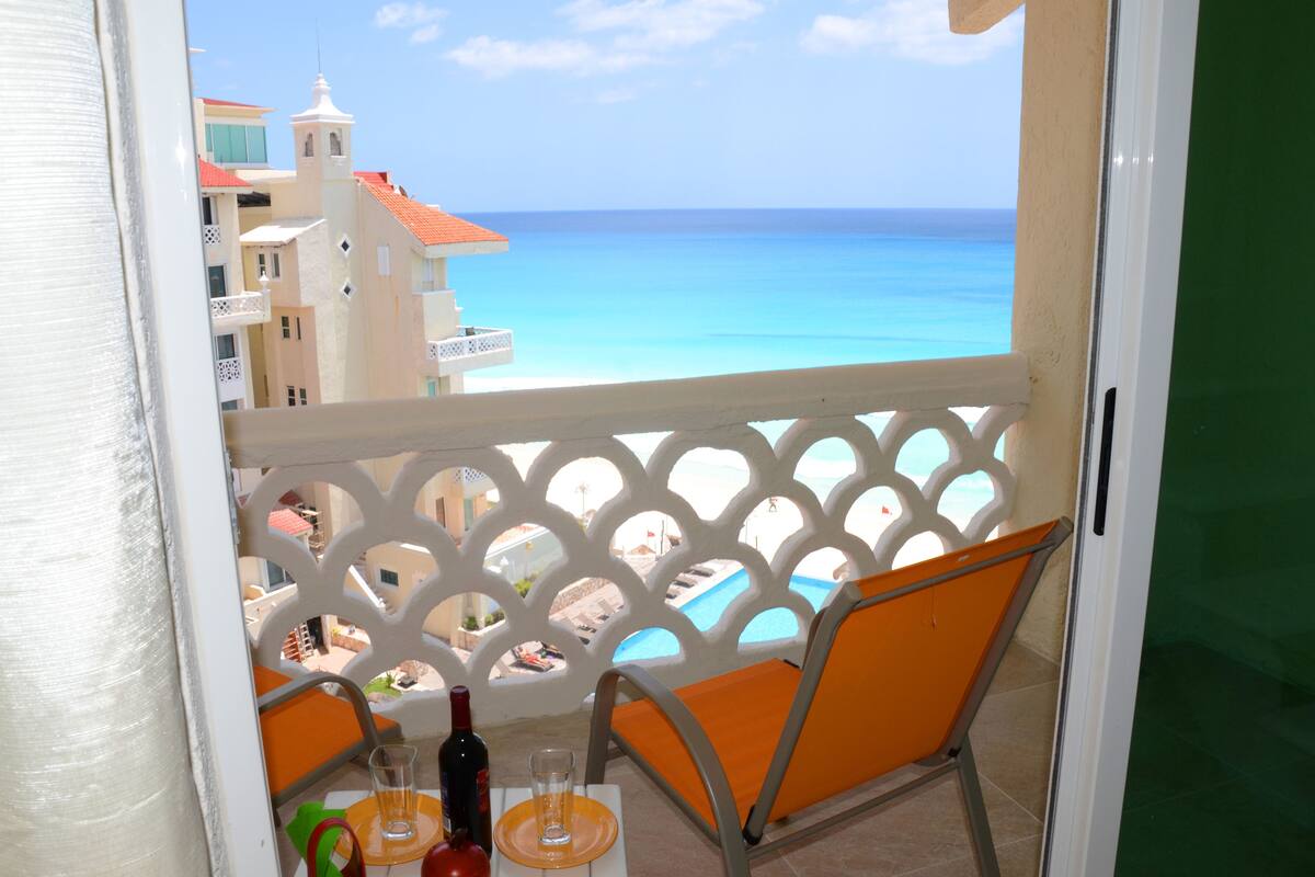 A balcony with a view of the ocean and buildings. There is a table with a bottle and glasses.