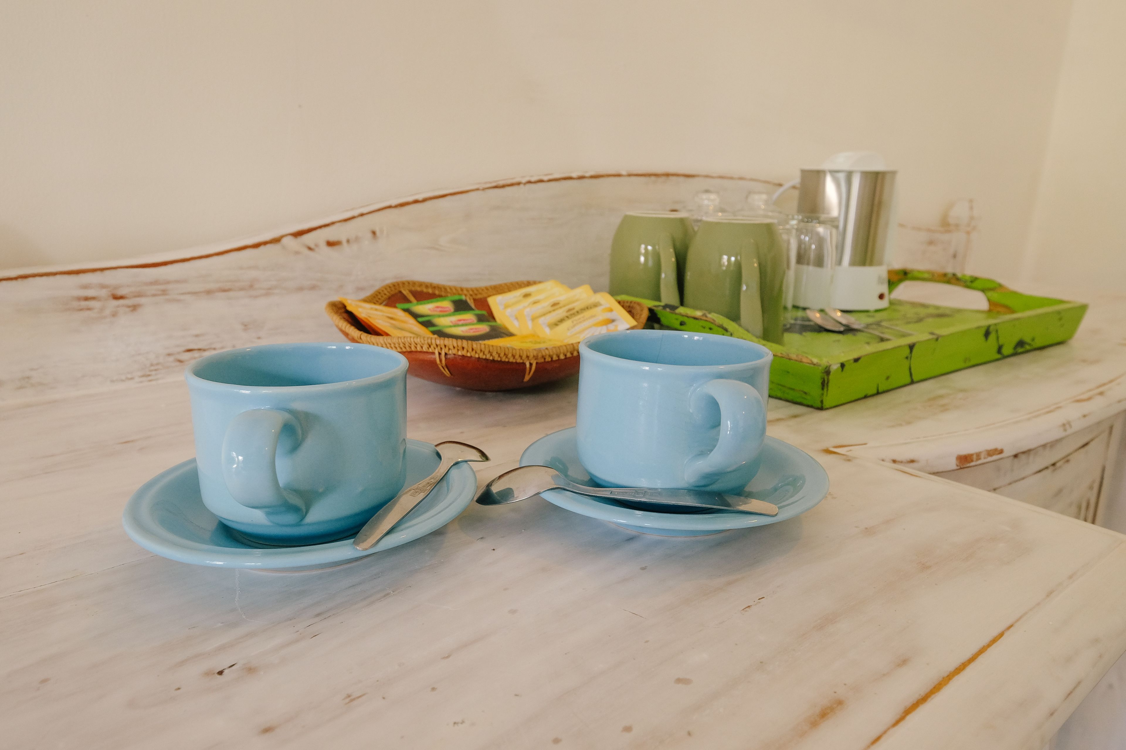 A table with two blue cups and saucers, a wicker basket with snacks, and a green tray with a teapot and cups.
