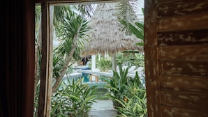 Visão através de uma porta para uma área de piscina com estrutura de telhado de palha, cercada por plantas tropicais e cadeiras de descanso.