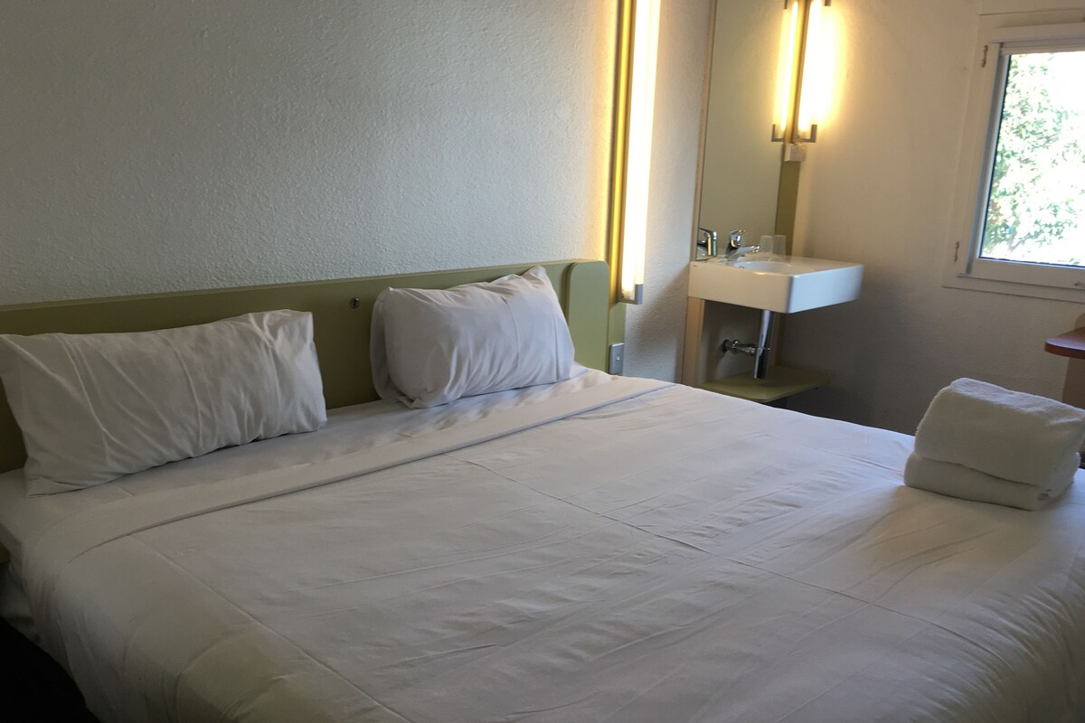 Une chambre d’hôtel comprenant un lit, une table de chevet, un miroir, une fenêtre et une salle de bain avec un lavabo.