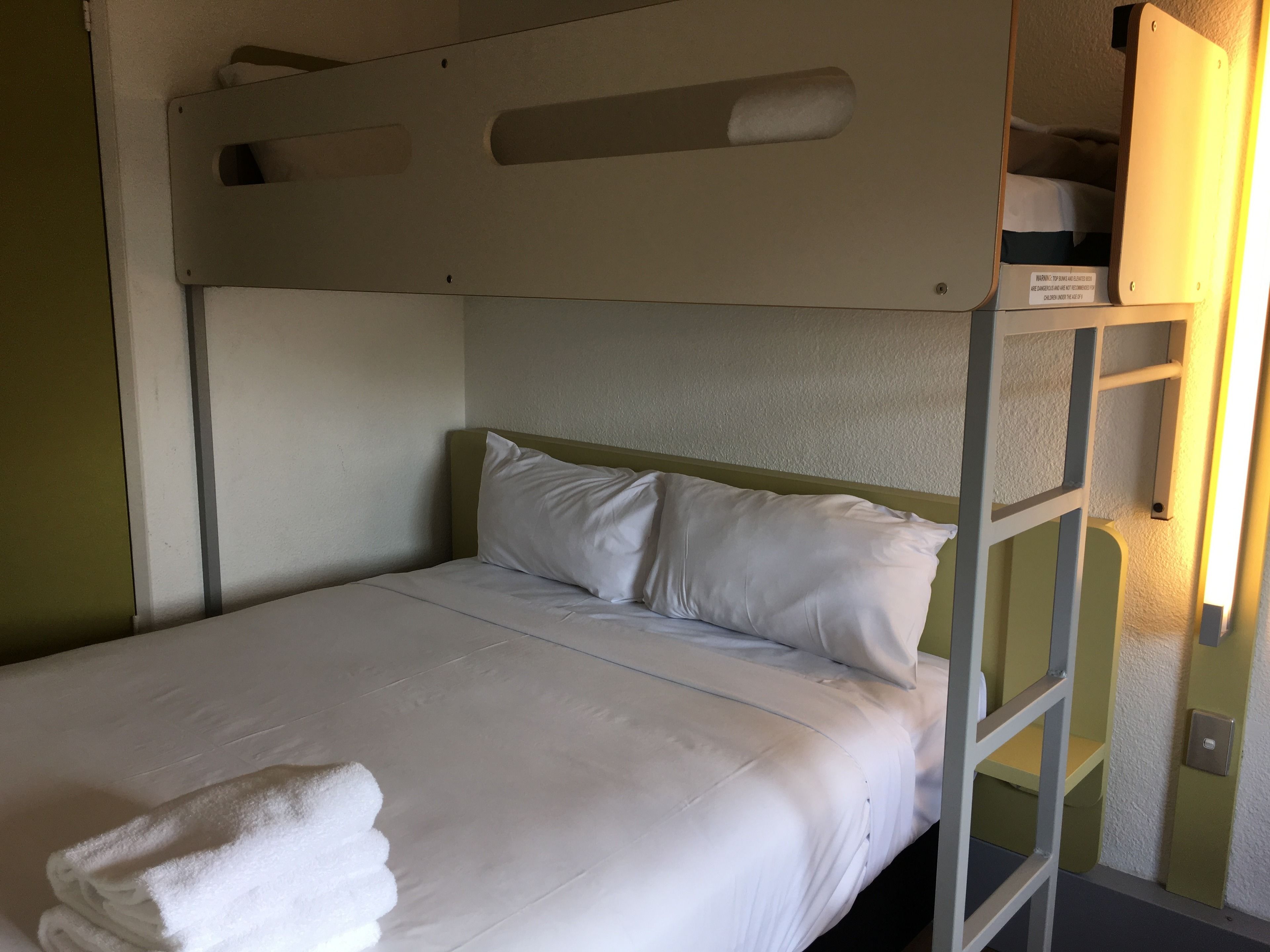 A single bed with white bedding and pillows, a folded white towel, and a bunk bed above.