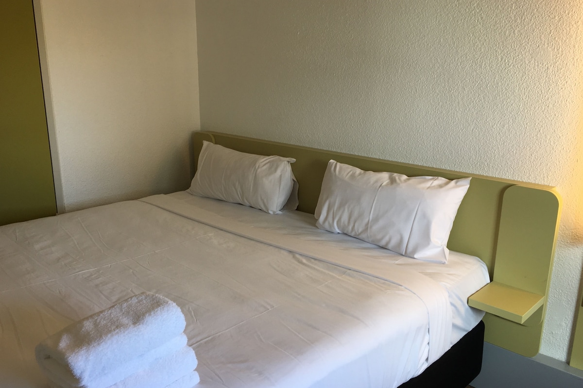 Un lit bien fait, avec du linge de lit blanc et des oreillers, une tête de lit verte et une table de chevet surmontée d’une lampe de chevet de couleur claire.