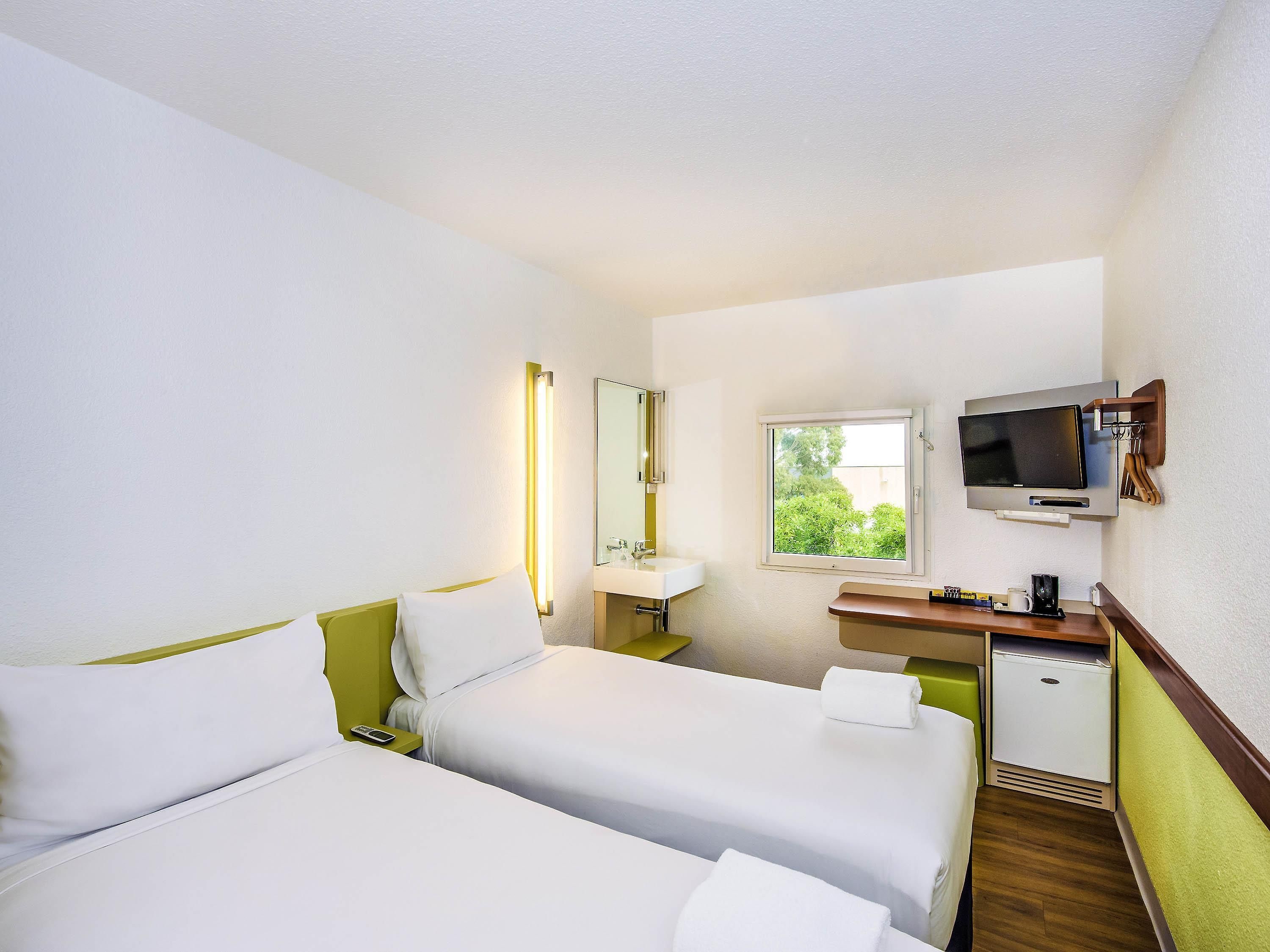 Ein Hotelzimmer mit zwei Betten, einem Fernseher, einem kleinen Schreibtisch mit Spiegel, einem Fenster und einem Kühlschrank.