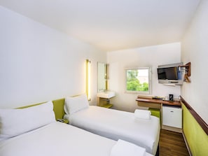 A hotel room with two beds, a TV, a small desk with a mirror, a window, and a refrigerator.