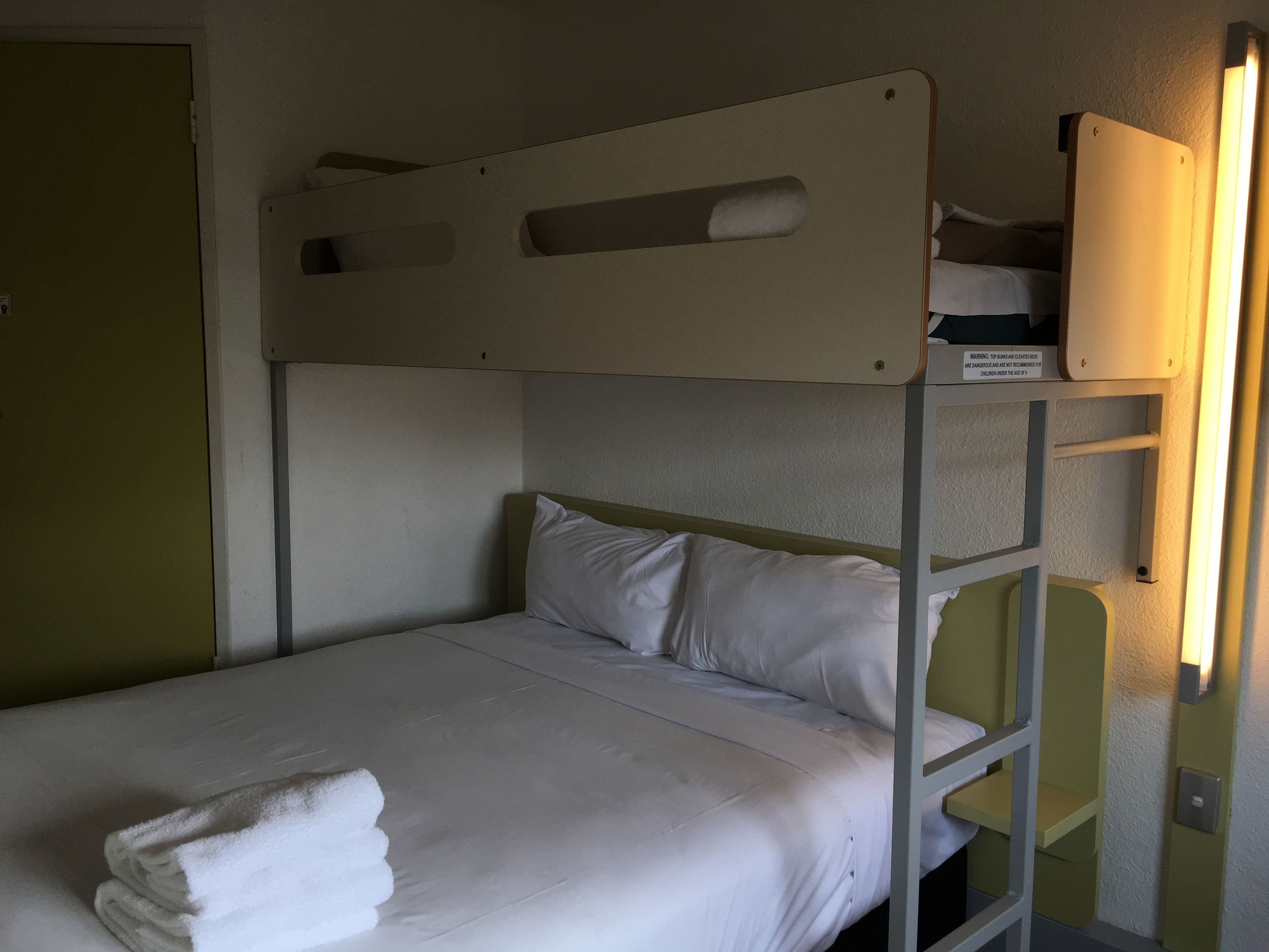 A bunk bed with a white mattress and pillows, a folded white towel on the lower bunk, and a wall-mounted shelf above the bed.
