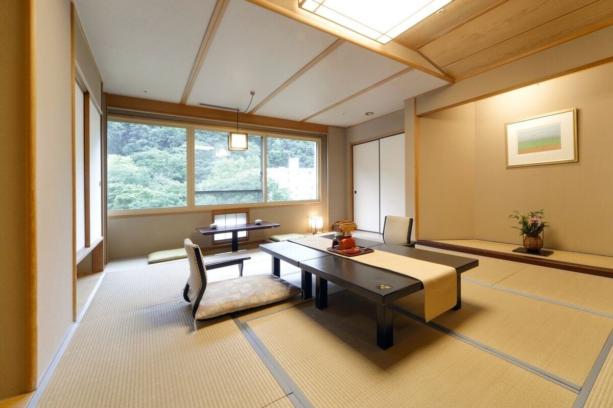 A traditional Japanese room with tatami flooring, a low table, a chair, and a window with a view of greenery.