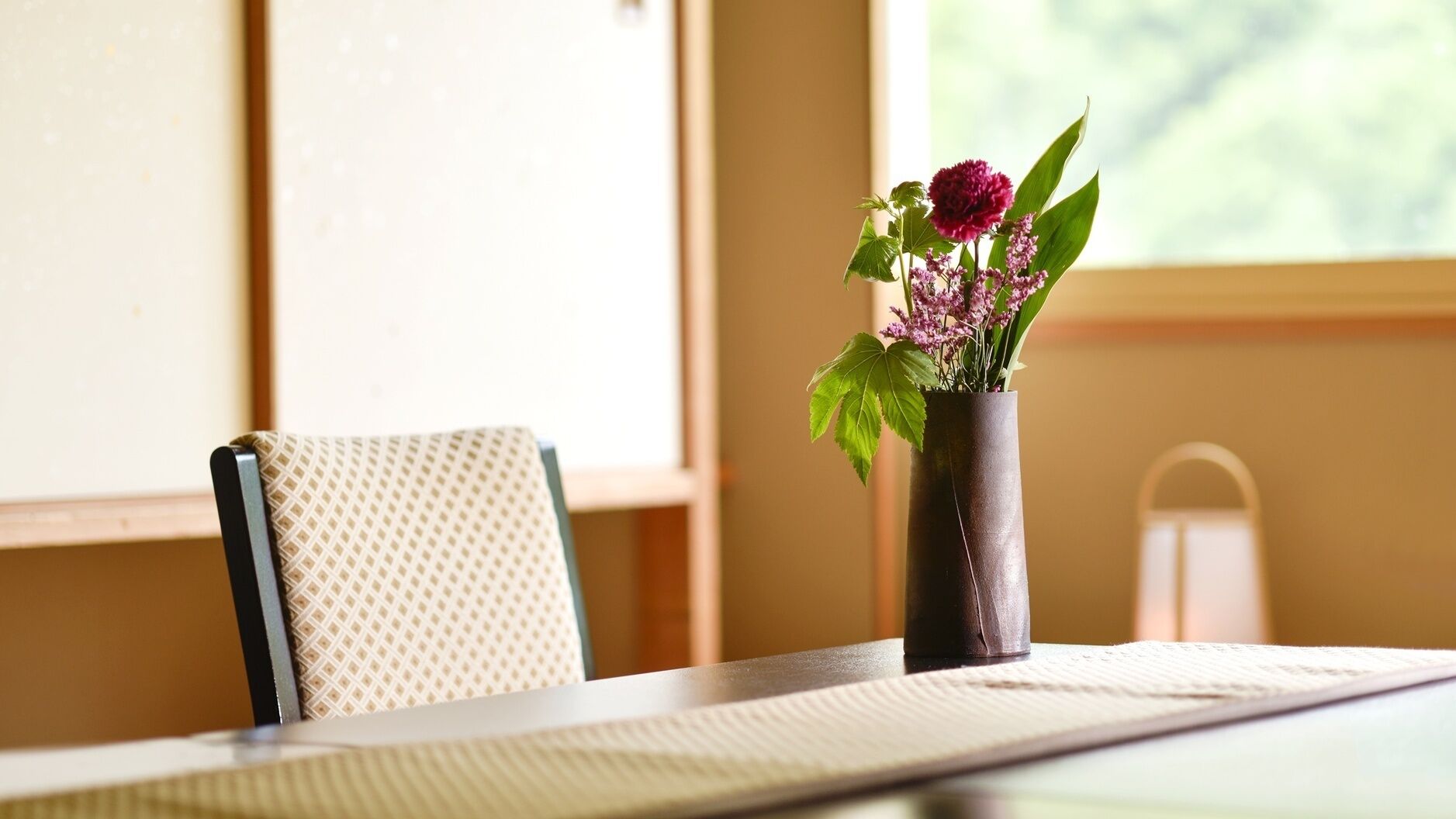 A table with a vase of flowers, a chair, and a window with a view.