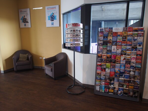 A reception area with a chair, a bulletin board with travel brochures, and a poster on the wall. - ibis budget Sydney Olympic Park (Sydney Olympic Park)