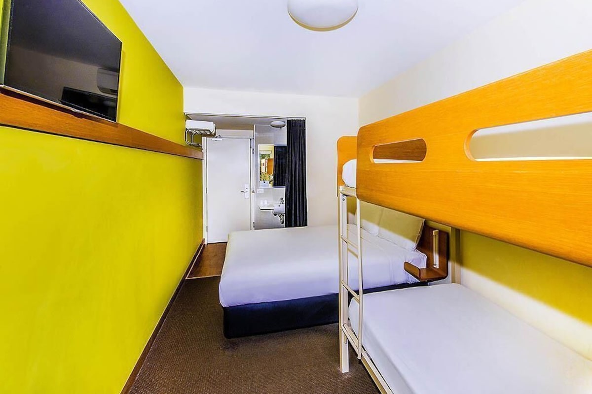 A compact hotel room with bunk beds, a small bedside table, a television, and a door leading to a bathroom.