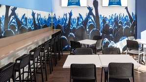 Een moderne hotellobby met een groot muurdecoratie van een concertpubliek, verschillende tafels en stoelen, en twee schermen waarop de naam van het hotel staat.