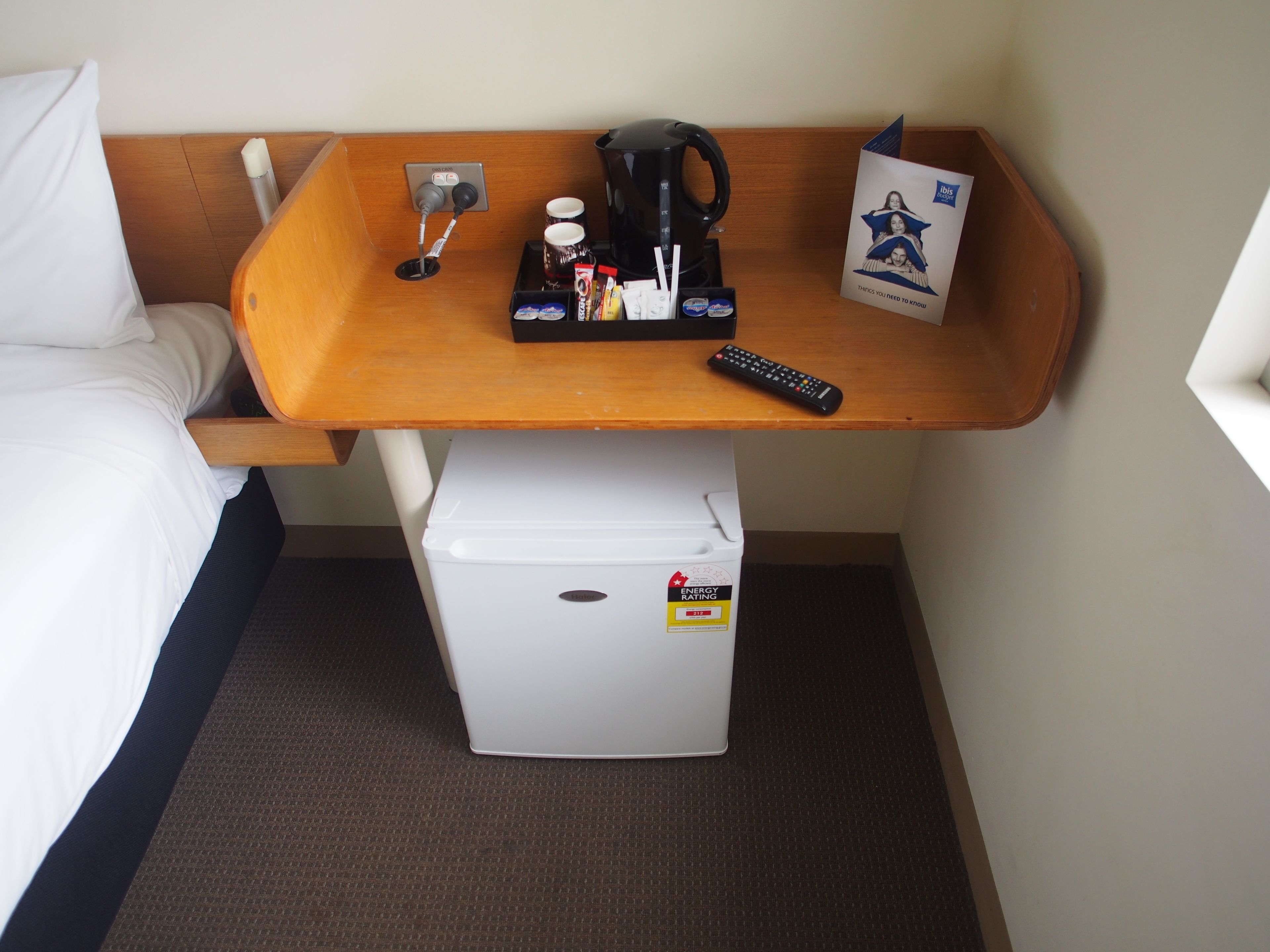 A compact hotel room with a bed, a small white refrigerator, a wooden side table with a coffee maker, a remote control, and a brochure.