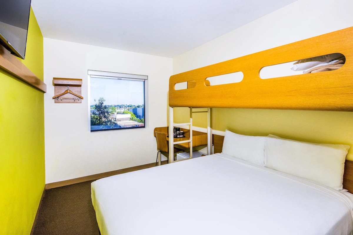 Une chambre d’hôtel avec un lit superposé, un lit simple, une fenêtre donnant sur la ville, une chaise en bois et un porte-manteau.