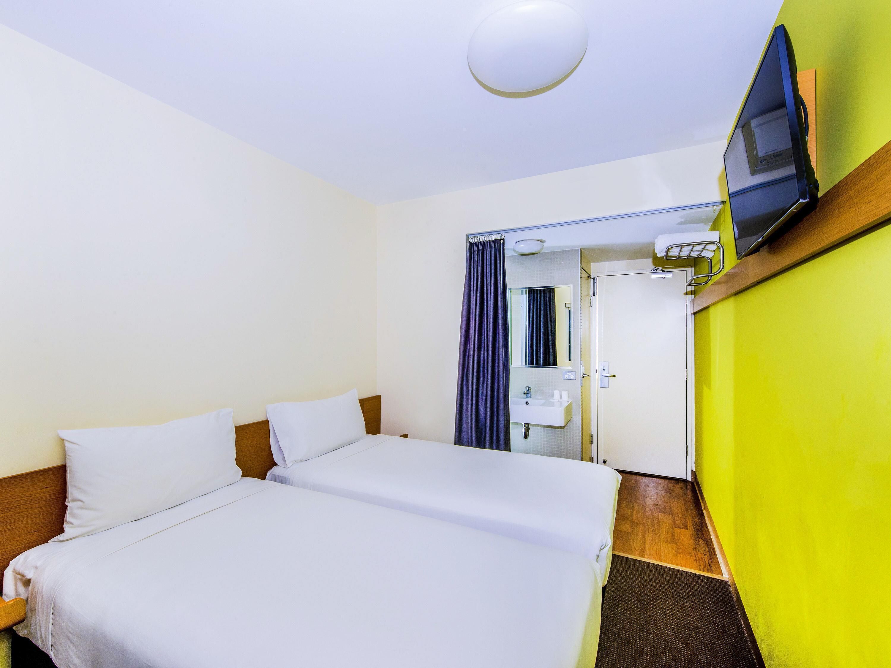 A hotel room with two beds, a bathroom with a sink and mirror, and a television mounted on the wall.