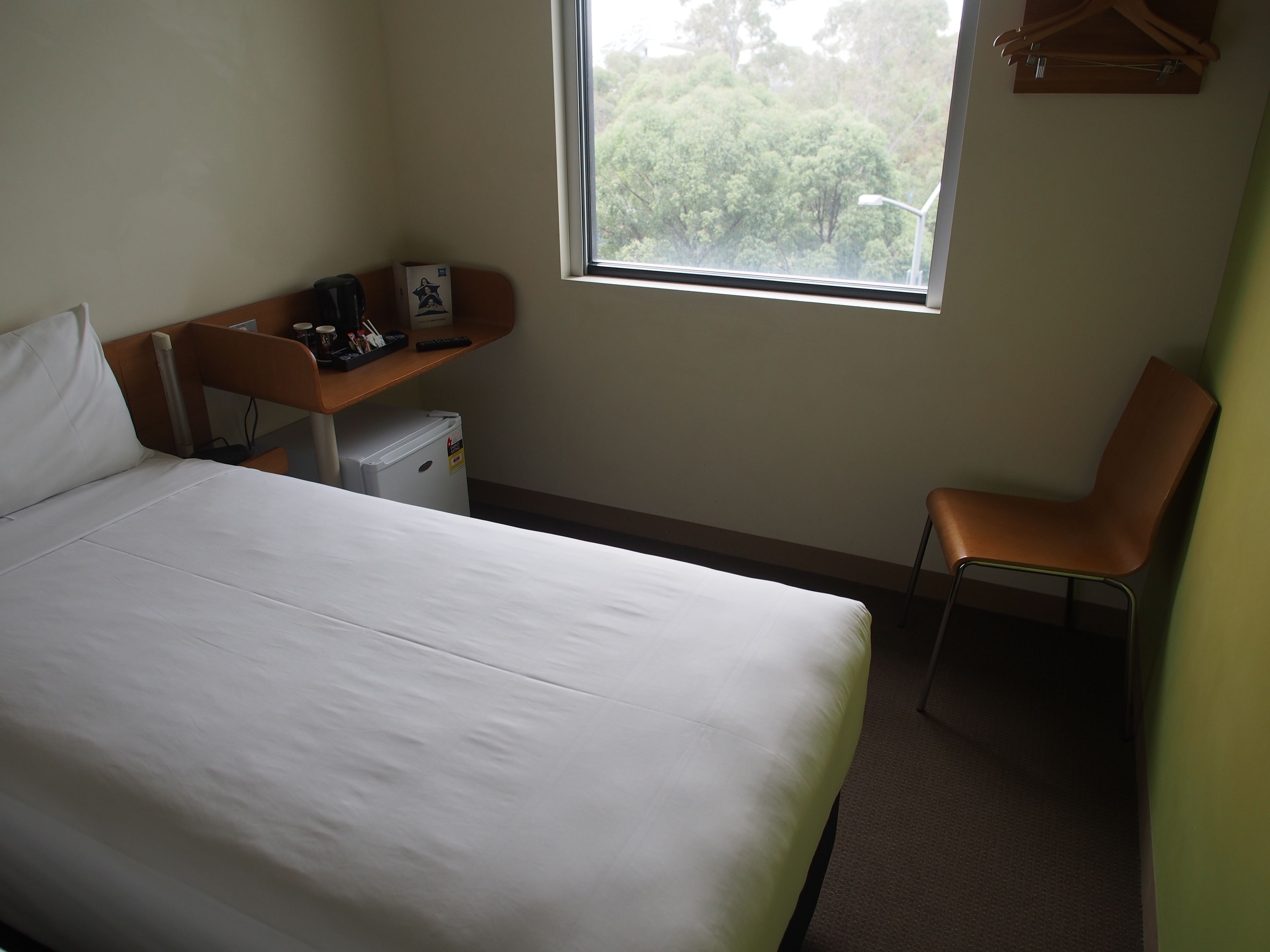 Ett hotellrum med en säng, ett skrivbord, en stol, ett litet kylskåp och ett fönster med utsikt över träd.