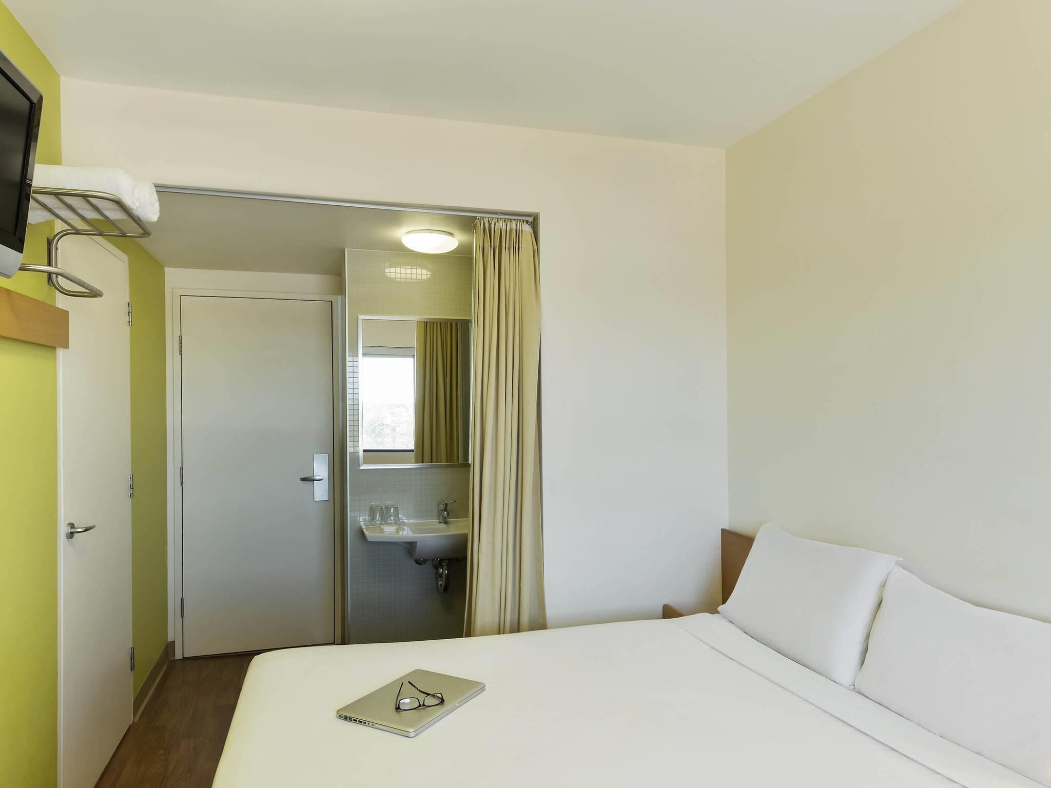 Habitación de hotel con cama, mesita de noche, espejo, baño con lavamanos y ducha.