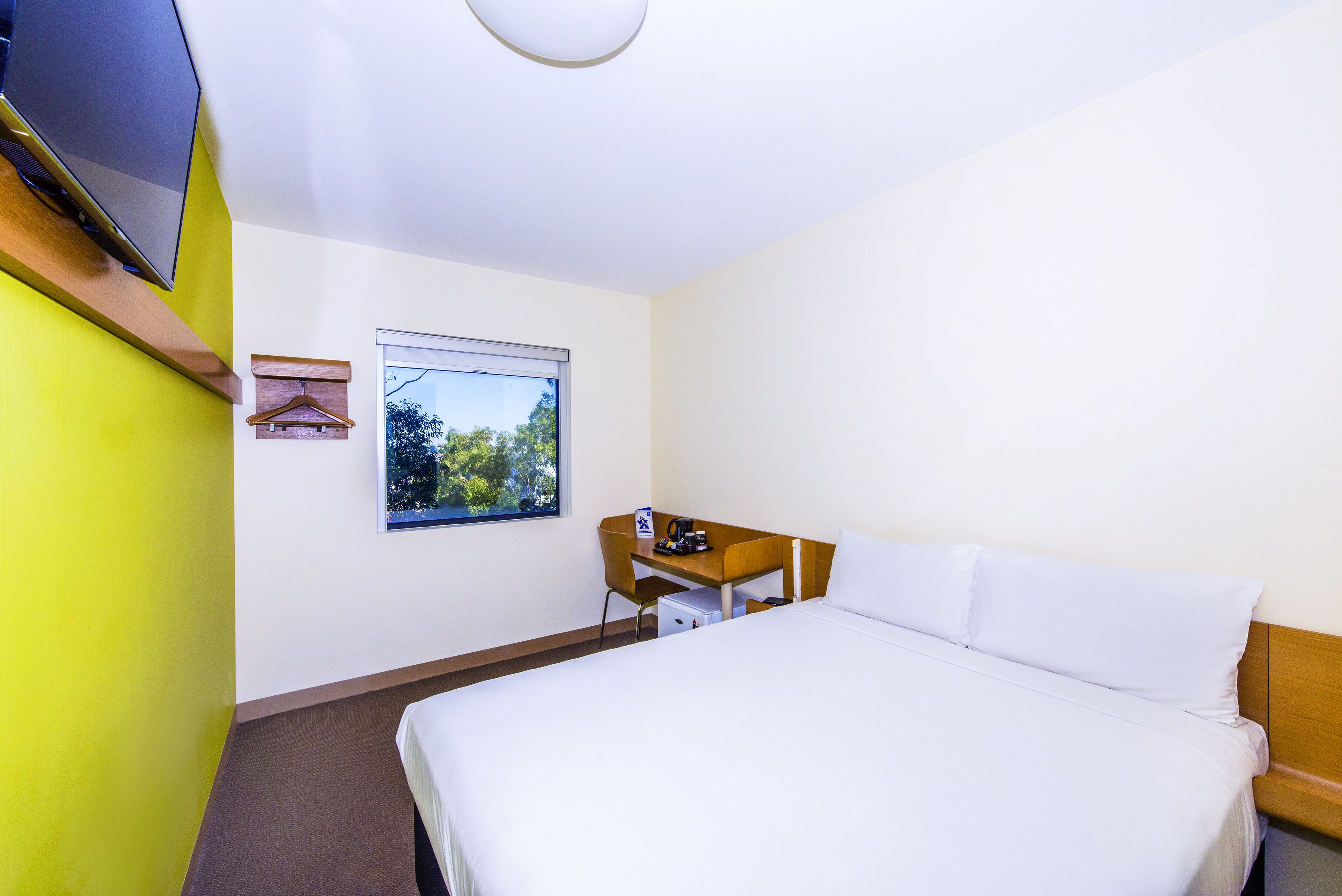 A hotel room with a bed, a desk, a chair, a TV, a window, and a coat rack.