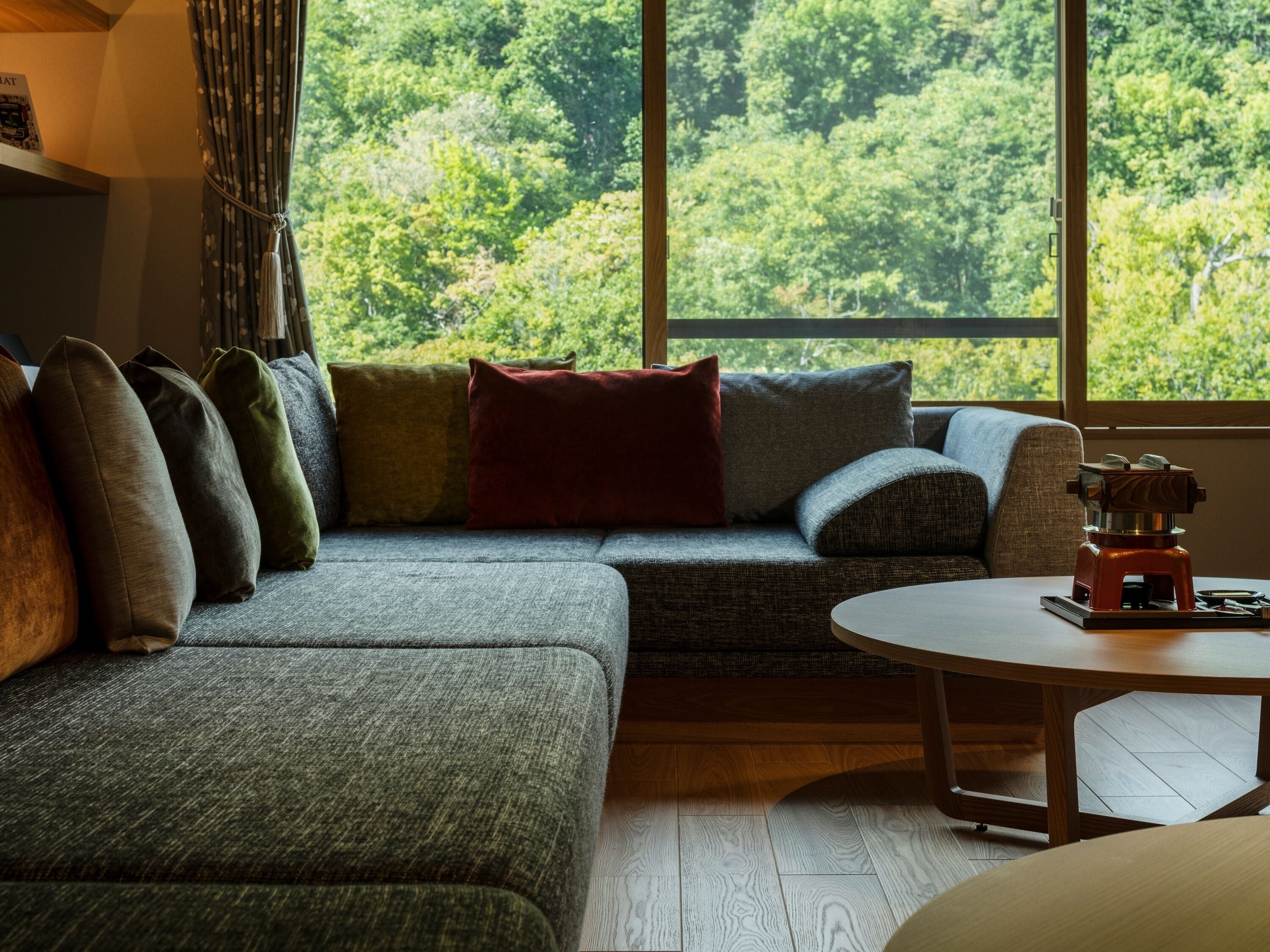 A cozy living room with a grey sofa, multiple cushions, a small round table, and a view of a dense forest through large windows.
