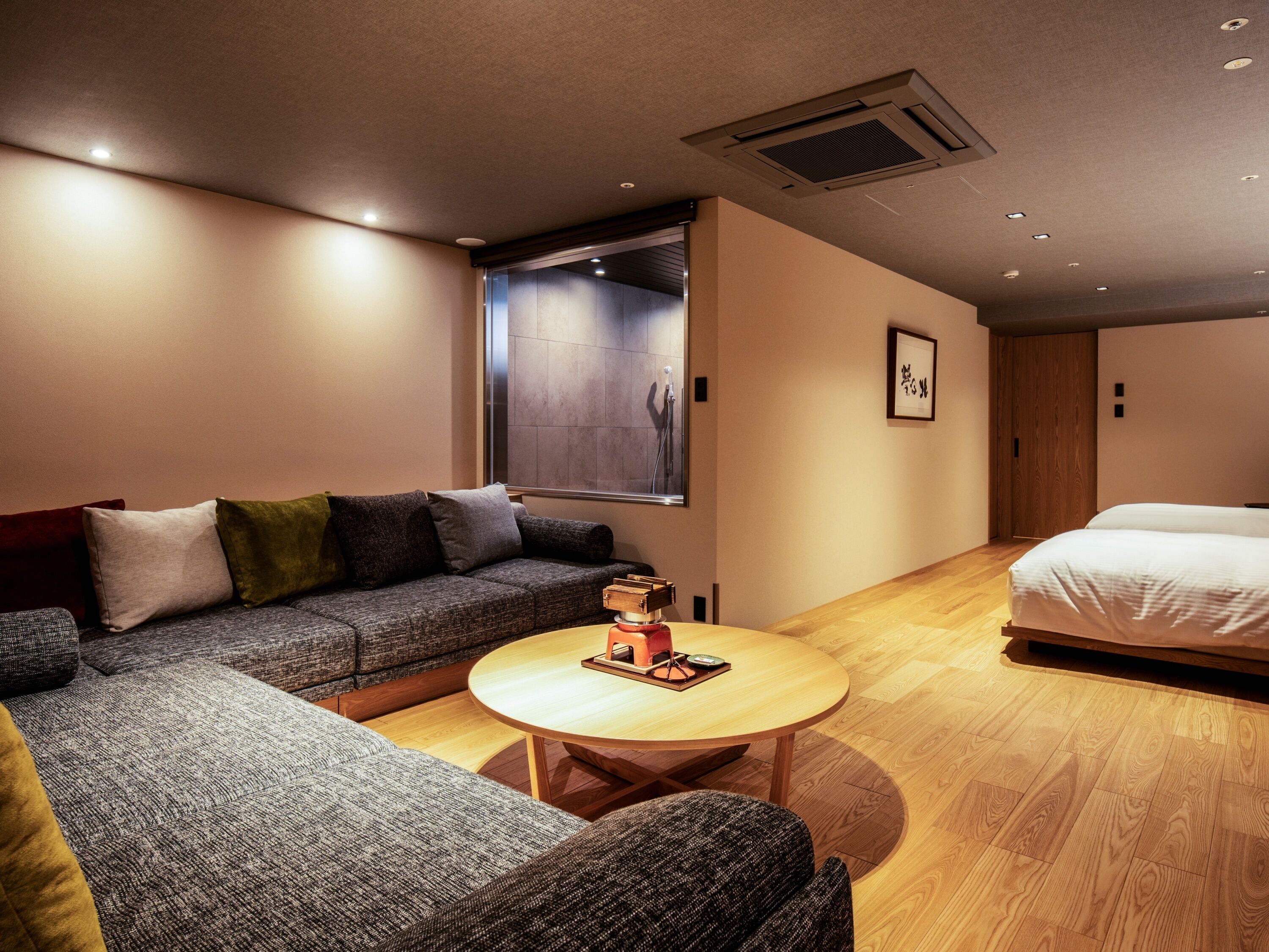 A modern hotel room with a sofa, a round wooden table, a bed, and a large window.