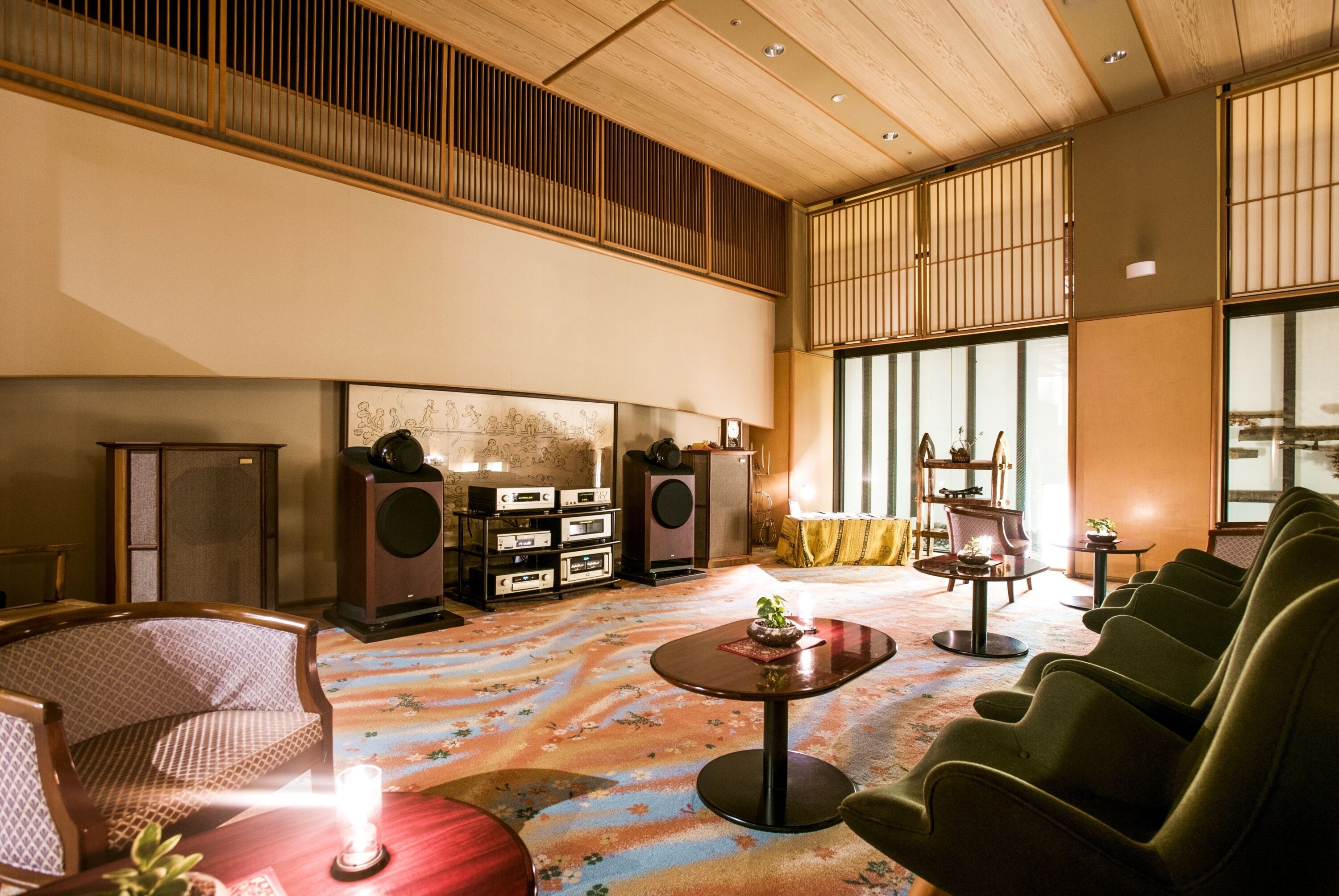 A spacious room with a high ceiling, a large window, a traditional Japanese-style room divider, a seating area with green chairs, a small table with a plant, and a carpeted floor.