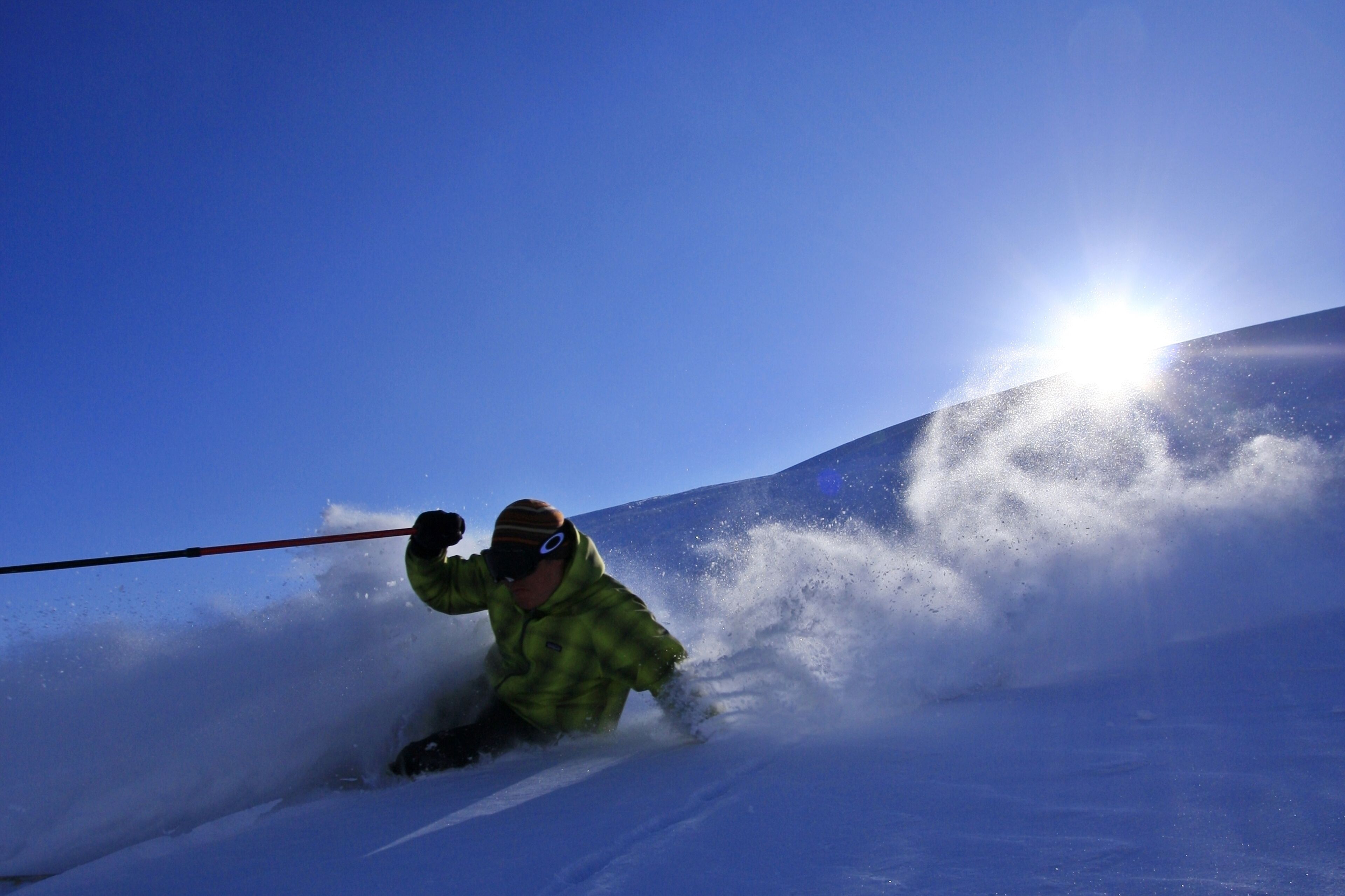A person skiing down a snowy slope with a clear blue sky and sun in the background.