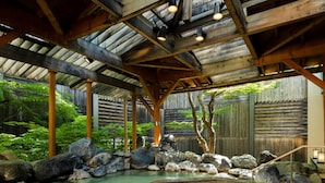 Un onsen japonés tradicional con estructura de madera, un manantial termal rodeado de rocas y una valla de madera al fondo.