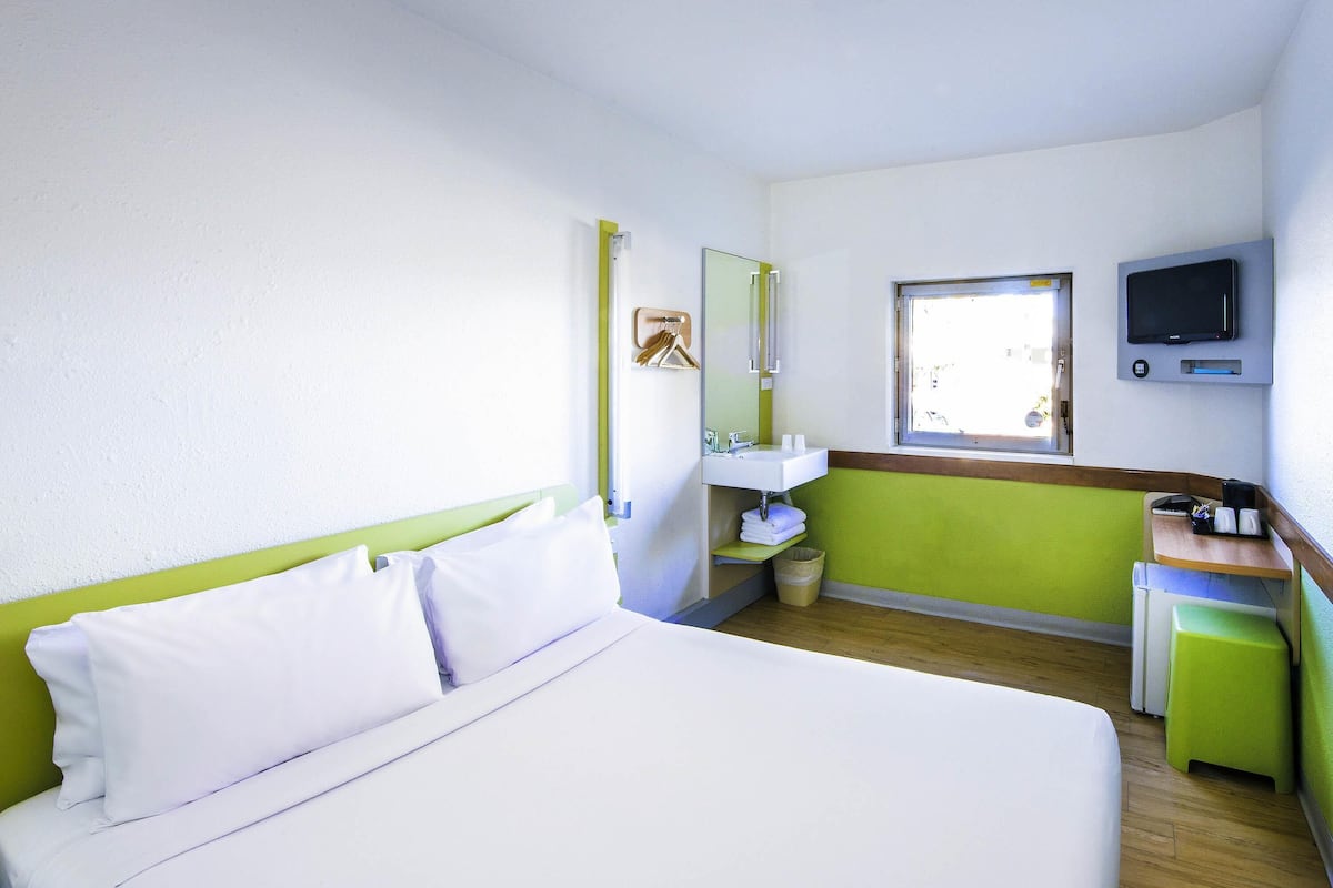 A hotel room with a bed, a sink, a mirror, a window, and a television.