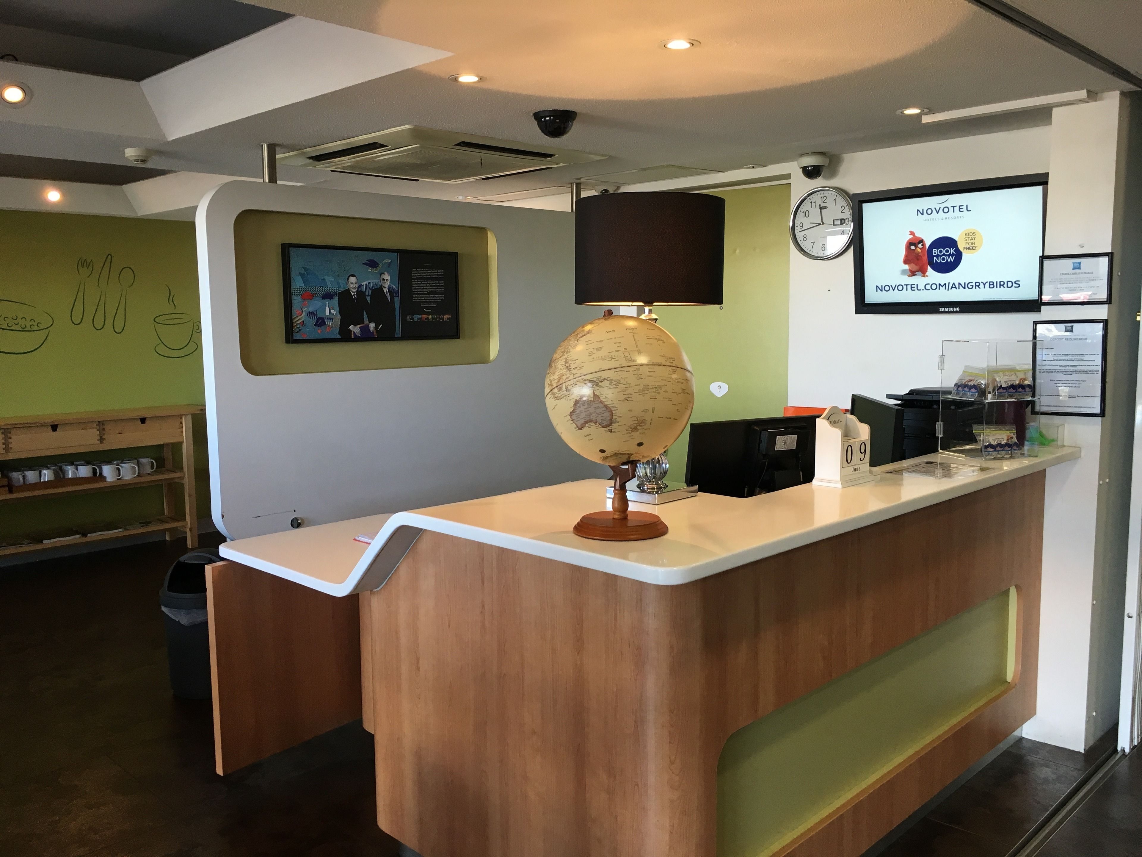 A reception desk with a globe lamp, a clock, and a digital screen displaying a Novotel advertisement.
