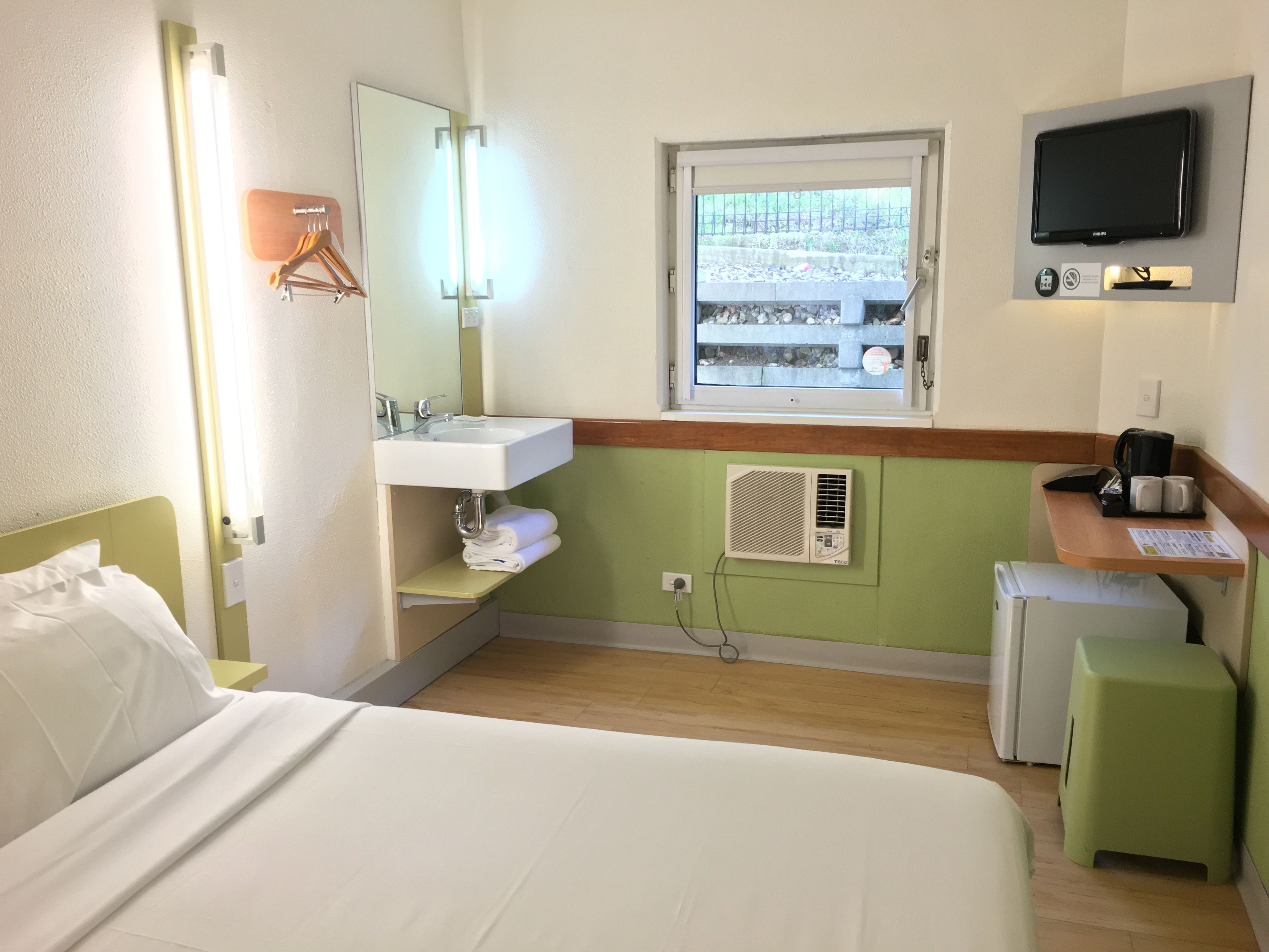 Habitación de hotel con cama, lavamanos, espejo, ventana con vistas, televisor y refrigerador pequeño.