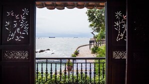A view through a wooden door with intricate cut-out designs, leading to a balcony overlooking a coastal area with a paved walkway and greenery. - Binh An Village Vung Tau (Ho Chi Minh City)