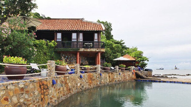 A stone wall with a wooden fence, a building with a tiled roof, and a body of water. - Binh An Village Vung Tau (Ho Chi Minh City)