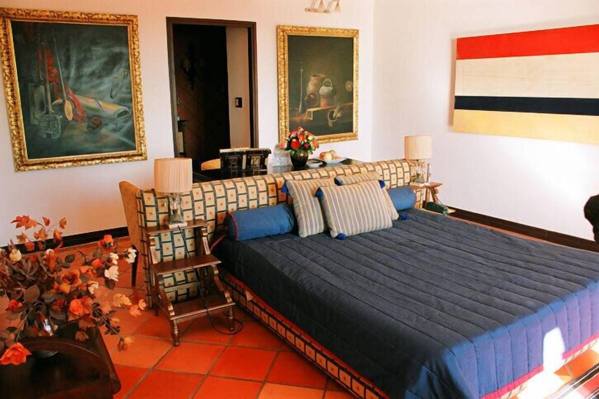 Un dormitorio con una cama grande, dos cuadros en las paredes, un jarrón con flores y una mesita de noche con una lámpara.
