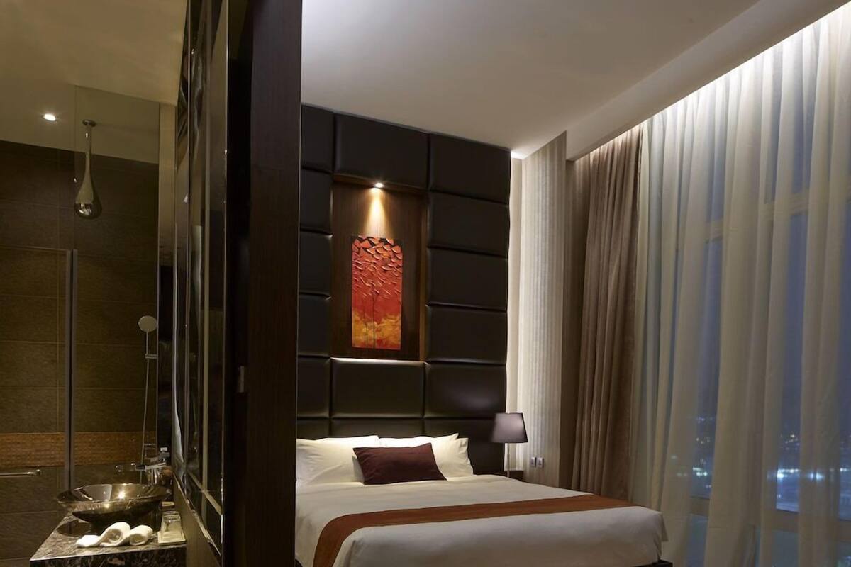 A modern hotel room with a large bed, a sitting area, a bathroom with a shower, and a city view.