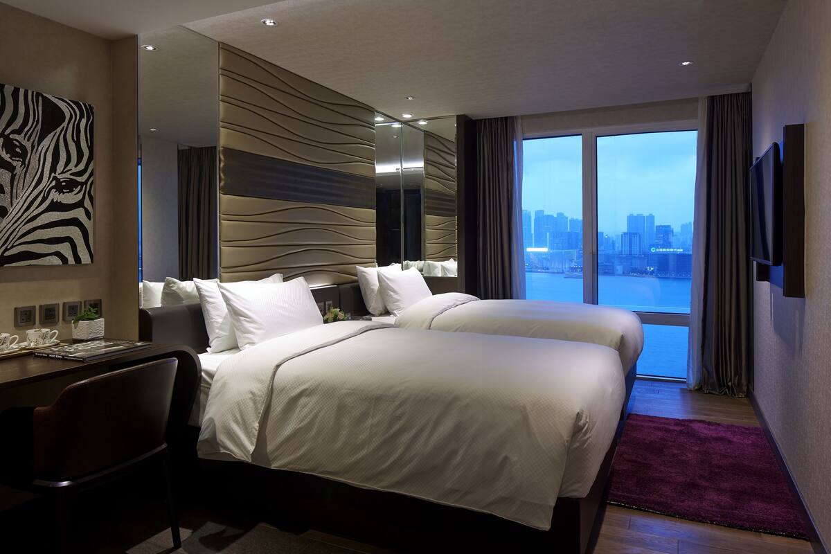A hotel room with two beds, a desk, and a large window offering a city view.