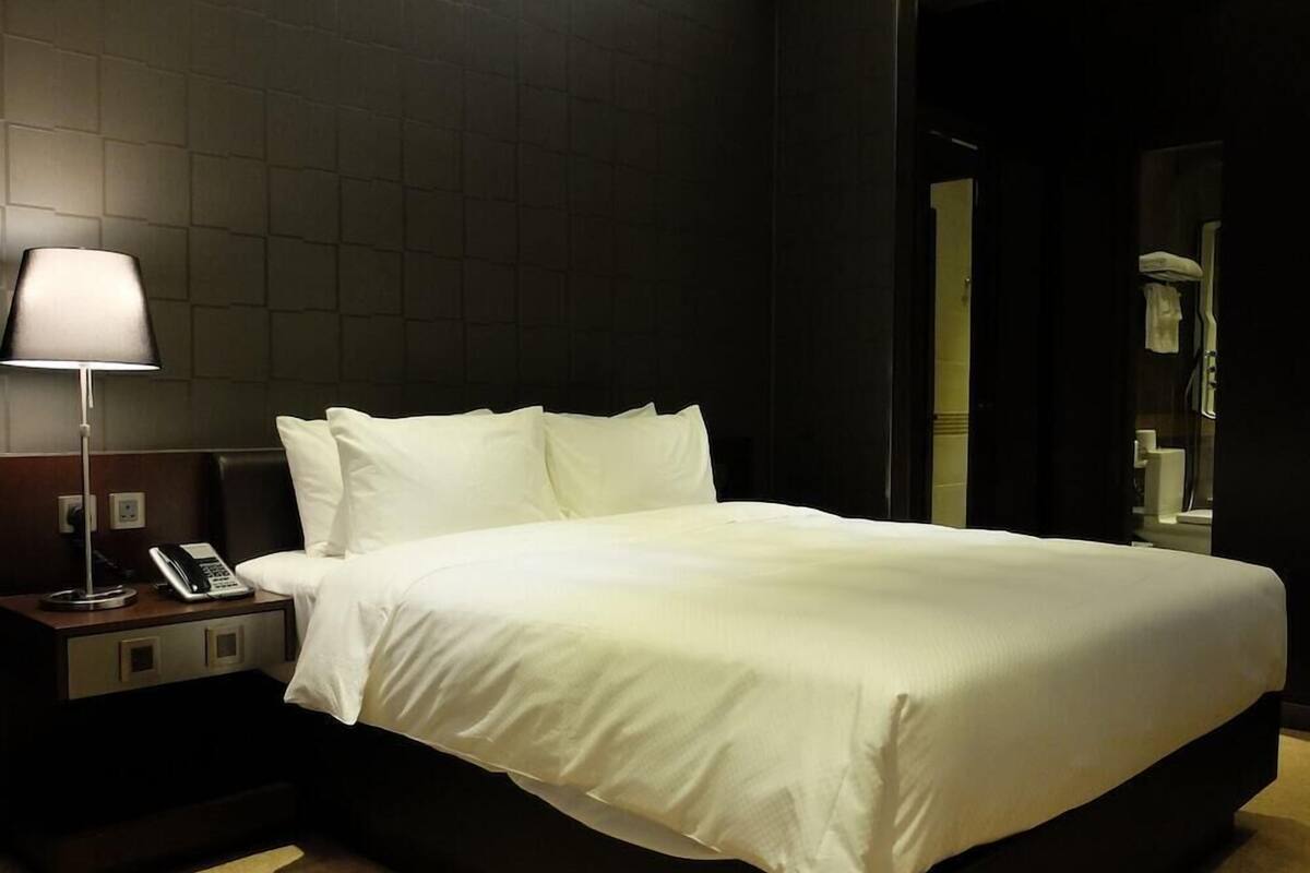 A hotel room with a large bed, a bedside table with a lamp and phone, and a dark wall.