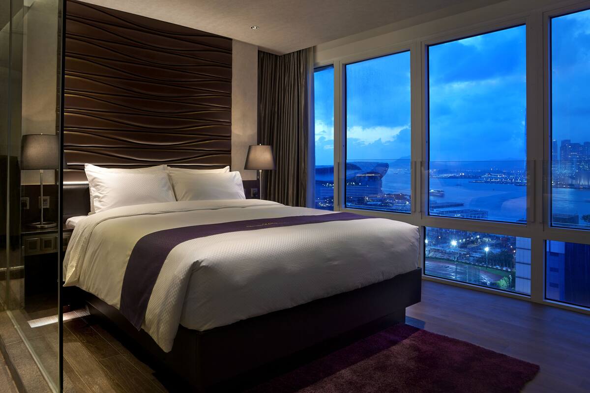 A modern hotel room with a large bed, a view of the cityscape, and a glass partition.