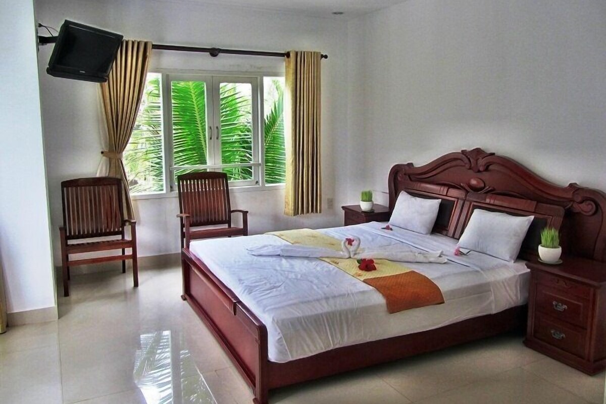 A hotel room with a bed, a TV mounted on the wall, a chair, a window with curtains, and a nightstand with a plant.