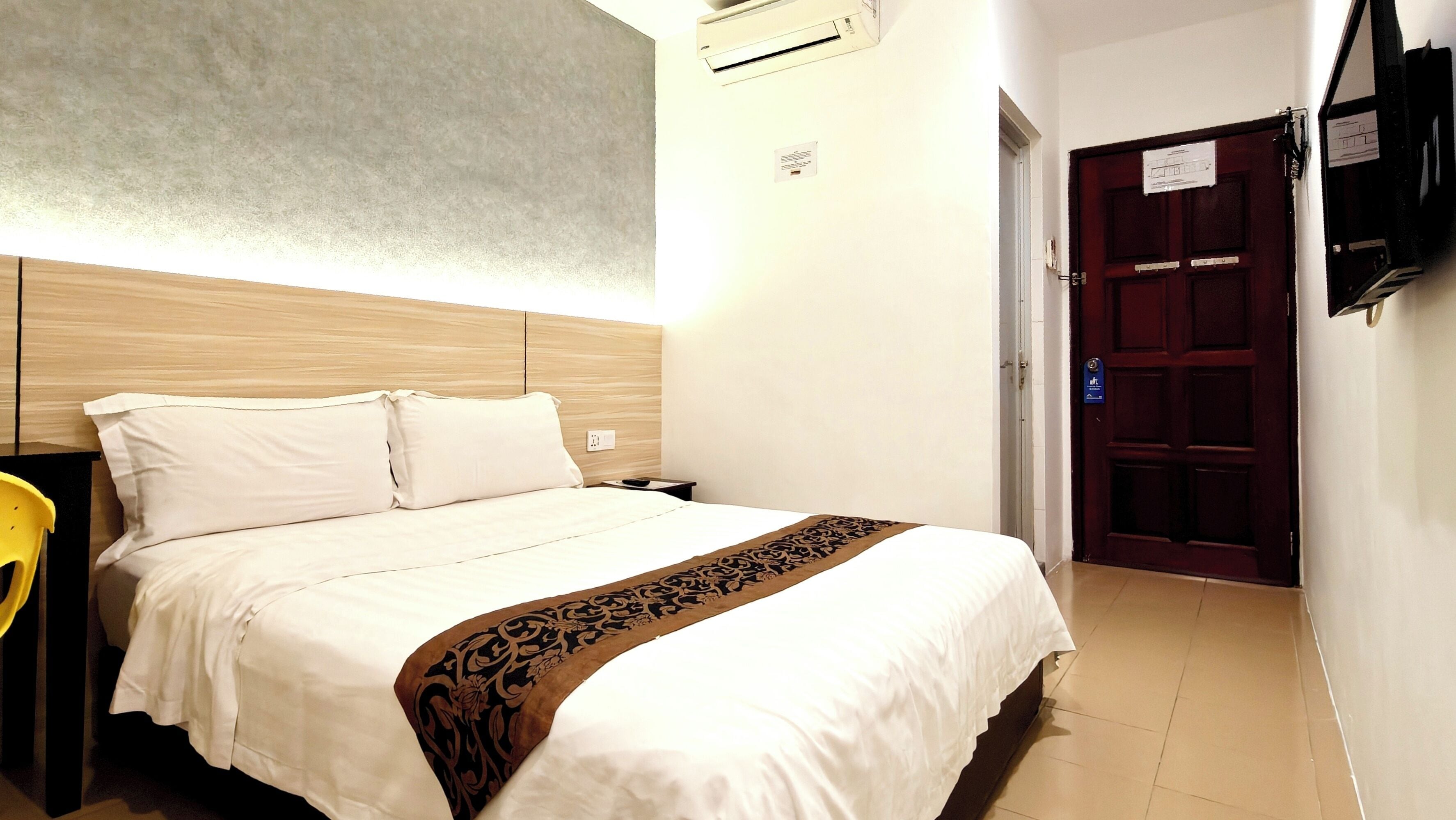 A hotel room with a bed, a nightstand, a chair, a wooden headboard, an air conditioning unit, a door, and a wall-mounted TV.