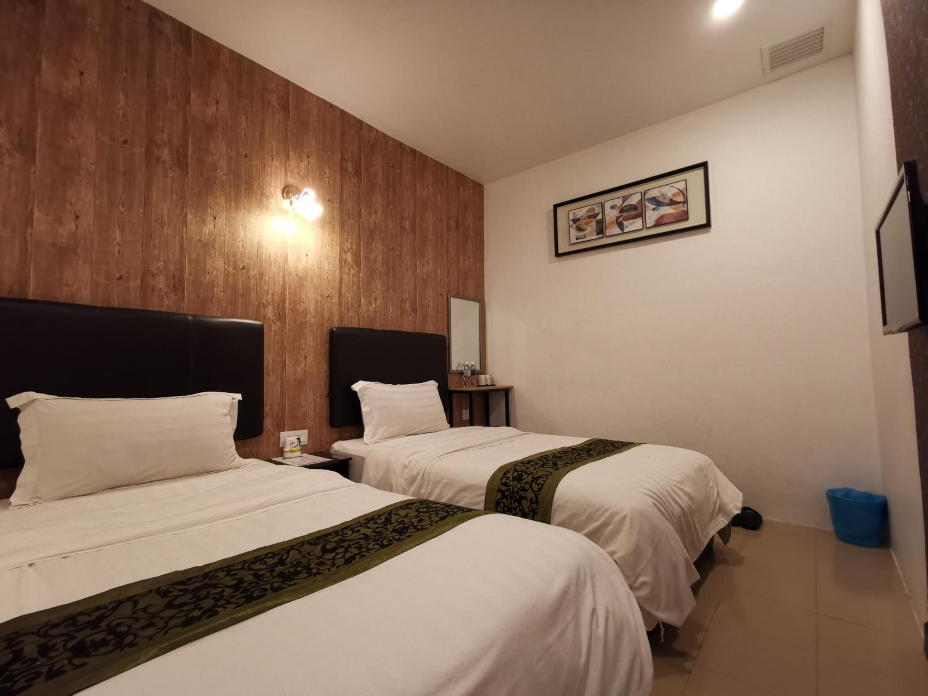 Habitación de hotel con dos camas, paneles de madera en la pared, una mesita de noche con lámpara, un cuadro enmarcado y un cubo de basura azul.