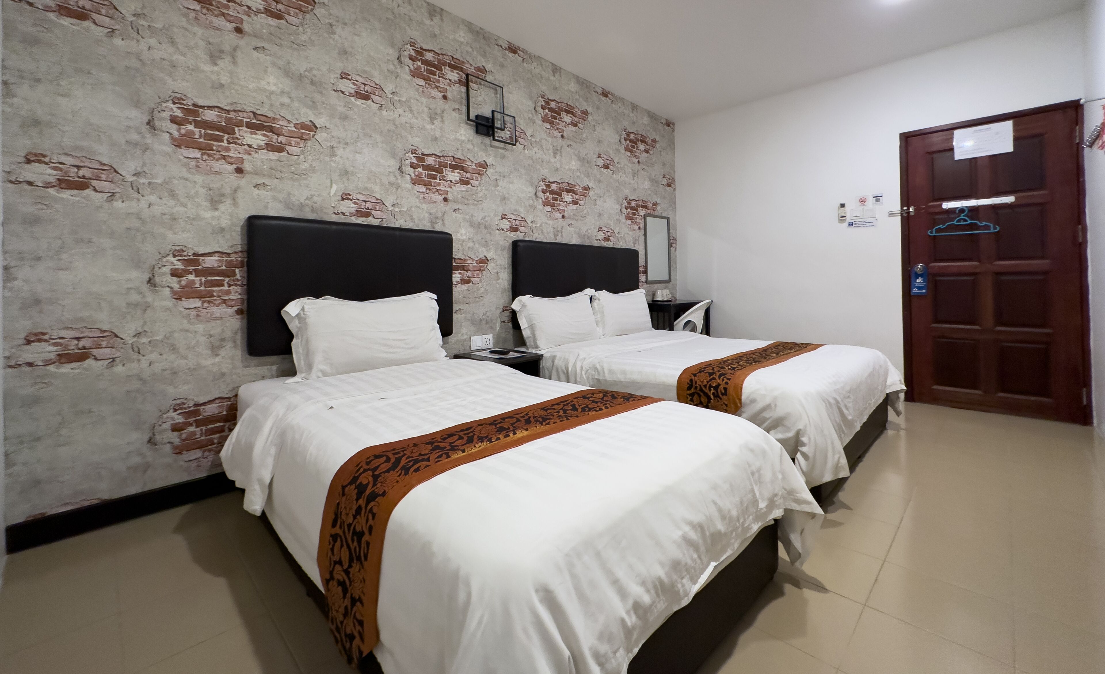A hotel room with two beds, a brick-patterned wall, a wooden door, and a tiled floor.