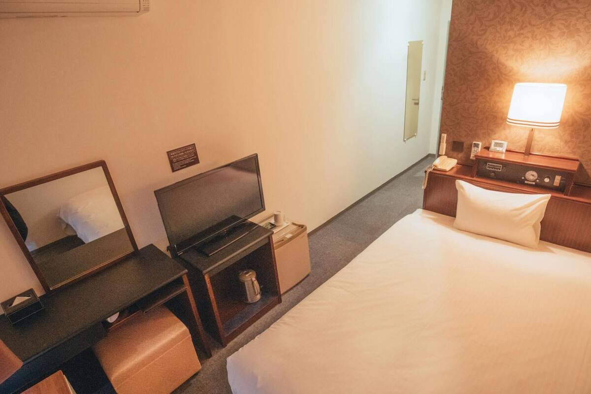 Ein Hotelzimmer mit einem Bett, einem Schreibtisch mit Computer, einem Sessel, einem Spiegel, einem Nachttisch mit Lampe und einem kleinen Safe.