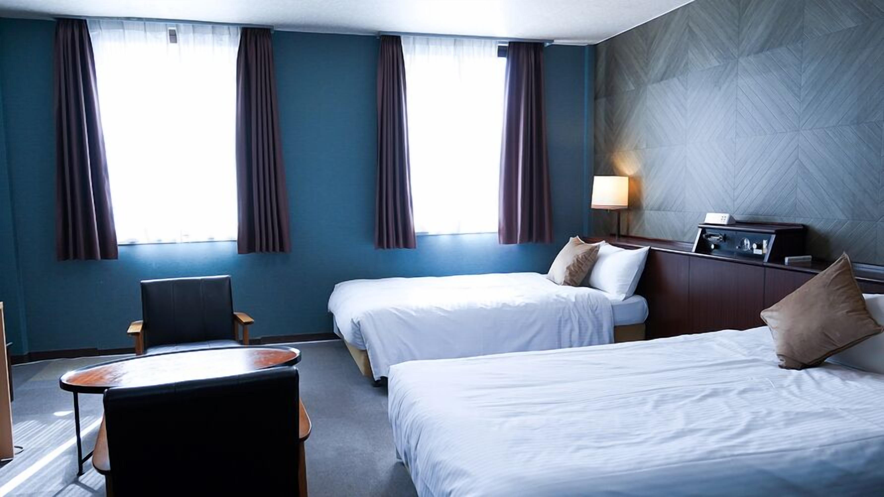 A hotel room with two beds, a desk, a chair, a nightstand, and a lamp.