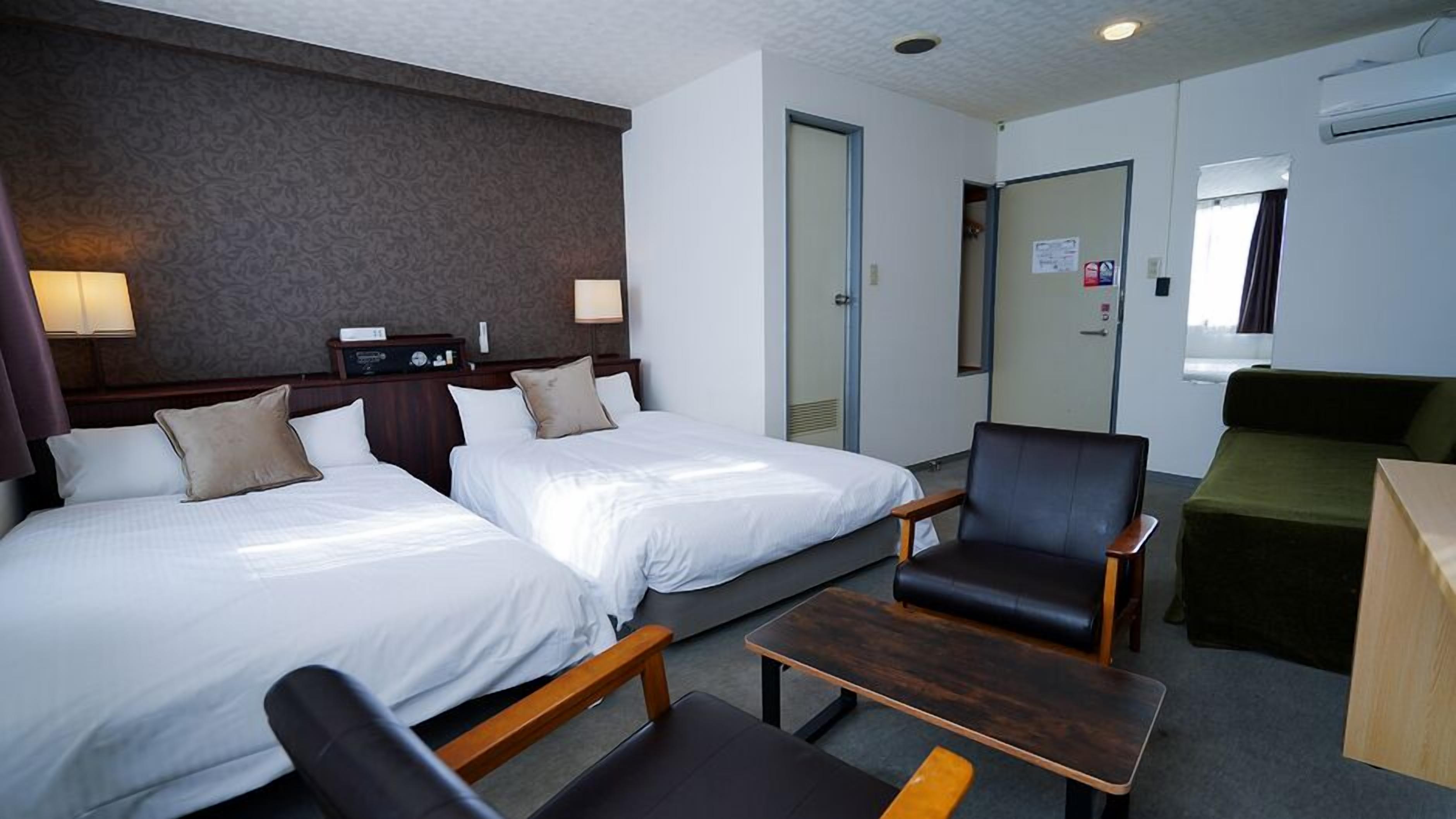A hotel room with two beds, a desk, a chair, a sofa, and a wall-mounted TV.