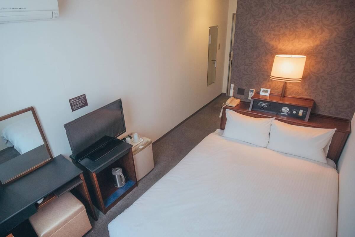 Ein Hotelzimmer mit einem Bett, einem Schreibtisch mit Stuhl, einem Nachttisch mit Lampe, einem Spiegel und einem kleinen Schrank.