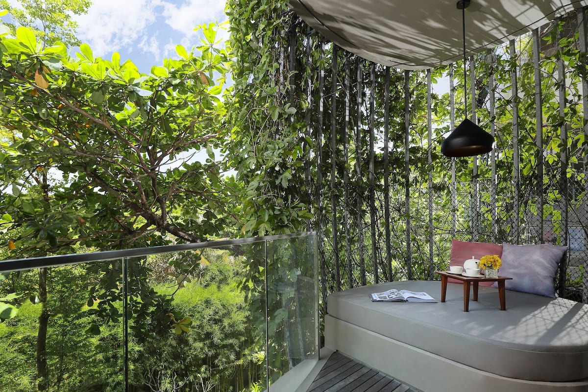 A balcony with a glass railing, a cushioned seating area, a hanging light fixture, and a view of dense foliage.
