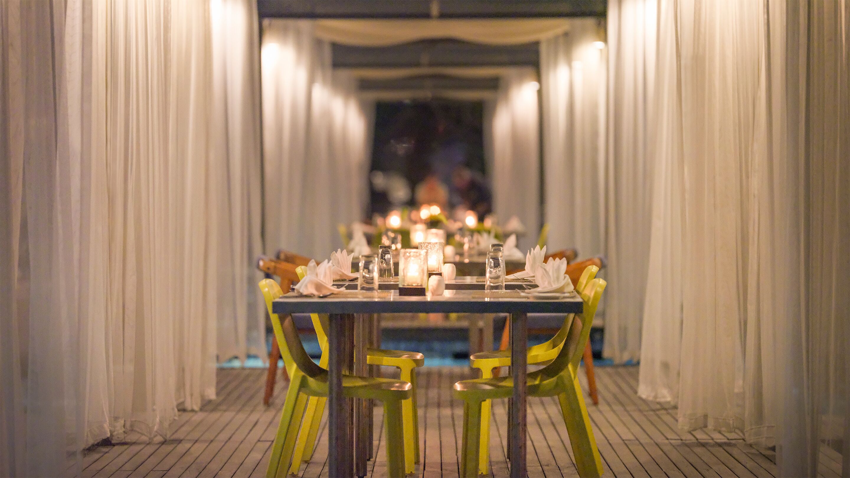 A dining area with a long table set for a meal, flanked by yellow chairs, and a pool in the background.