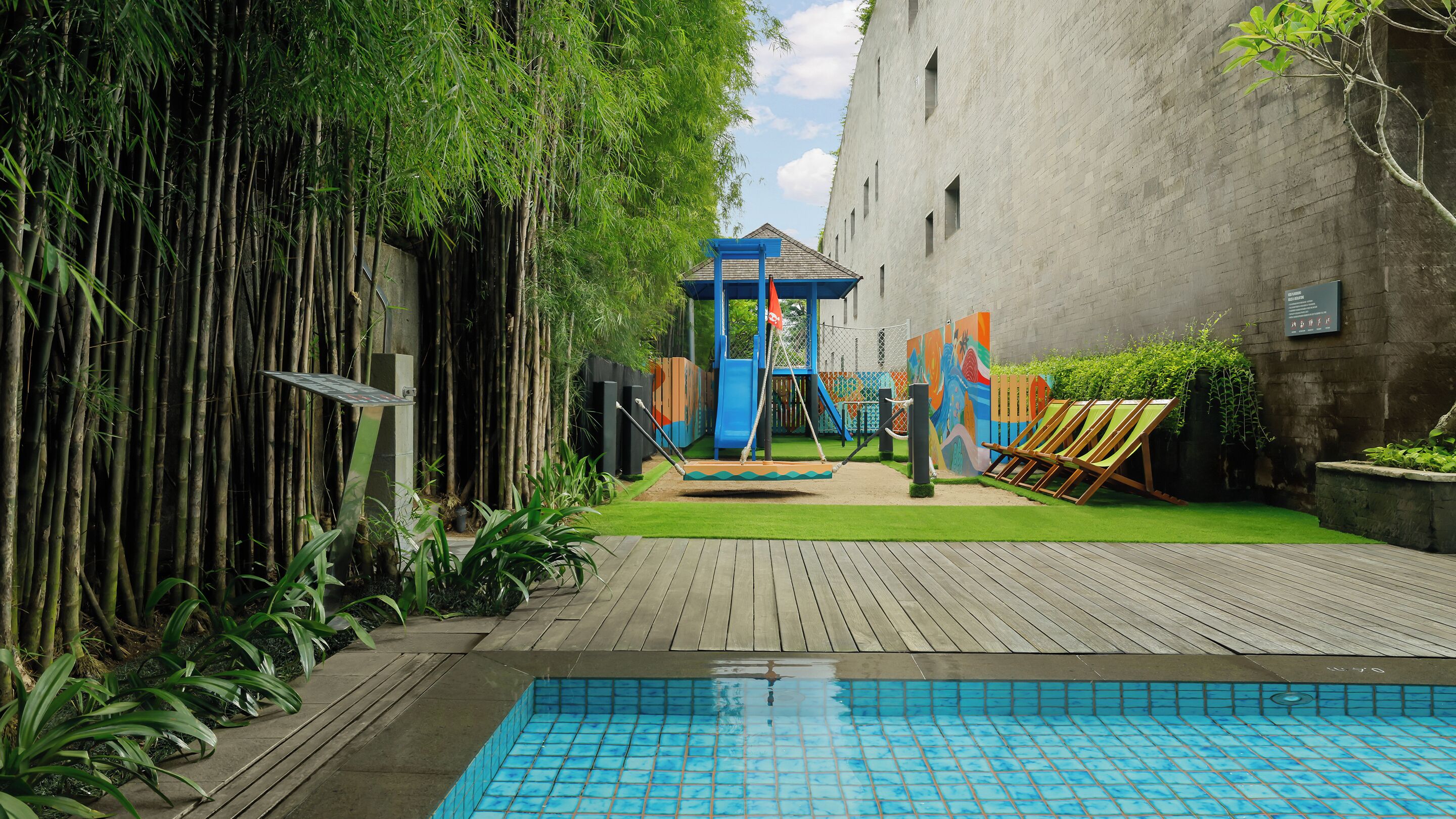 Una piscina con bordo in legno, un'area giochi per bambini con uno scivolo e piante di bambù.