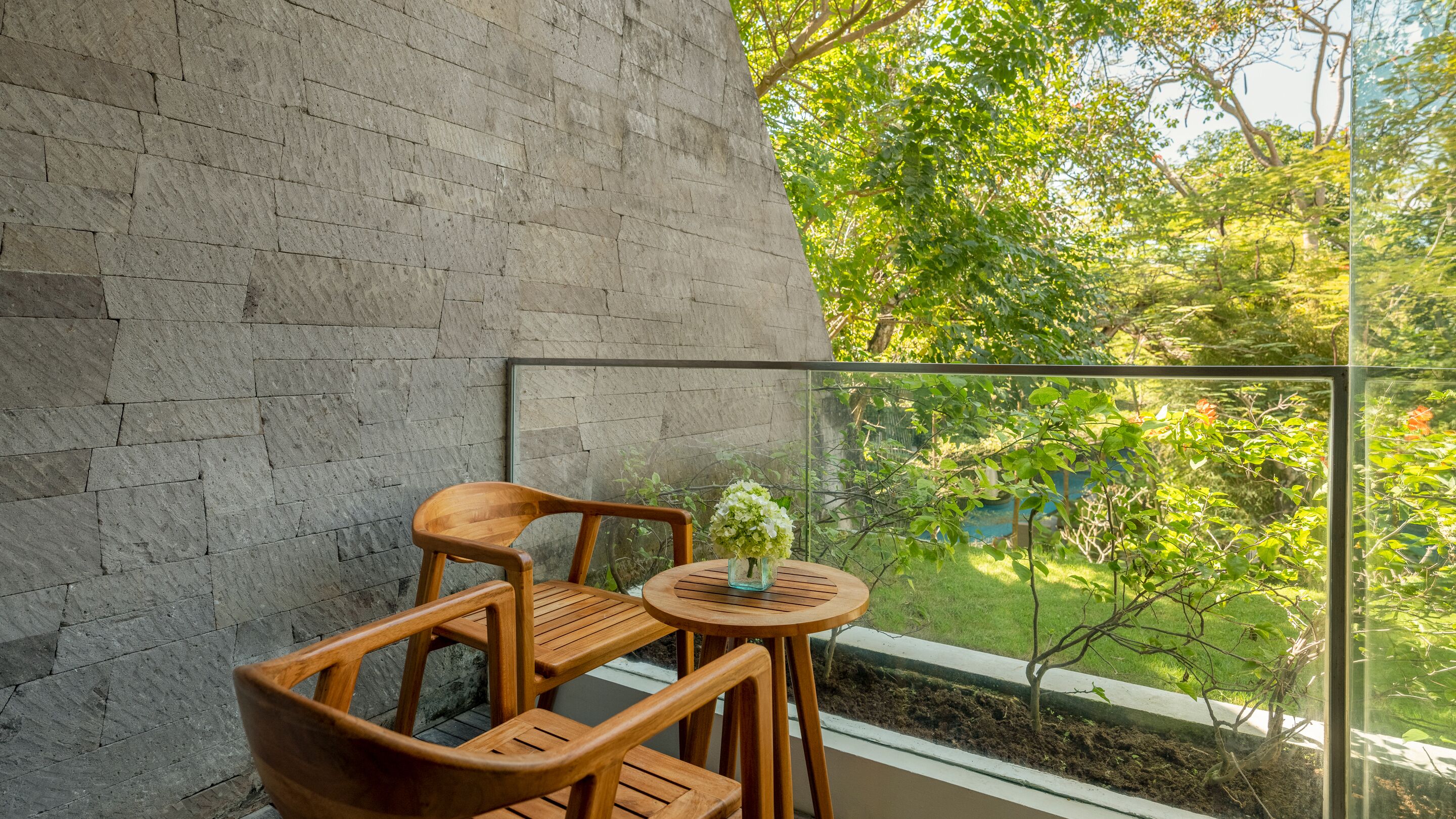 A balcony with a view of greenery, a wooden chair, a small table, and a vase with flowers.