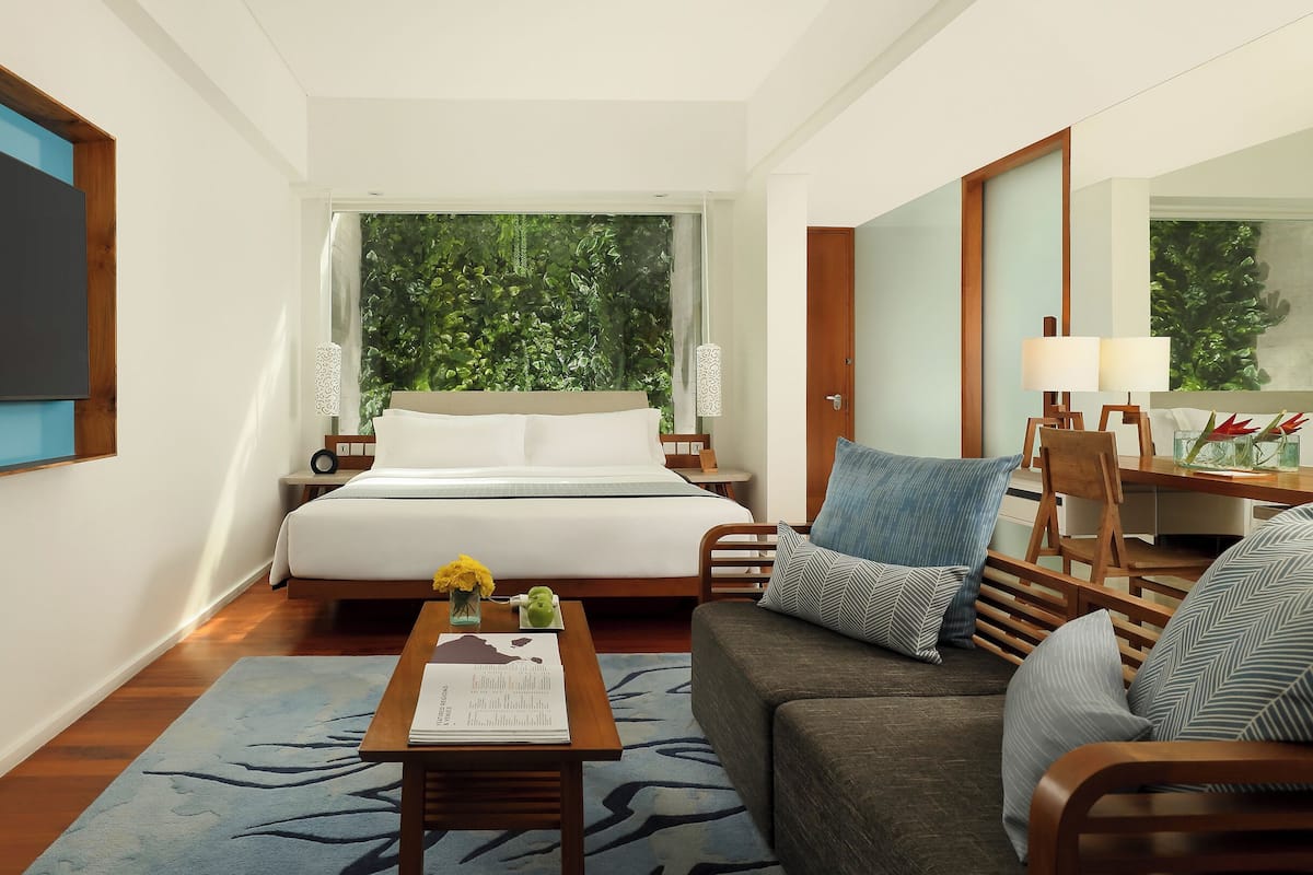 A hotel room with a bed, a sofa, a desk, and a large window with a view of greenery.